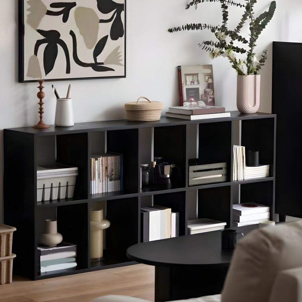 A modern grey 8-cube open bookcase positioned under a window with blinds. The minimalist wooden storage unit features a horizontal design with four open top shelves holding books and a camera, and four bottom shelves with woven storage baskets. A professional, clean-lined display for modern home offices.