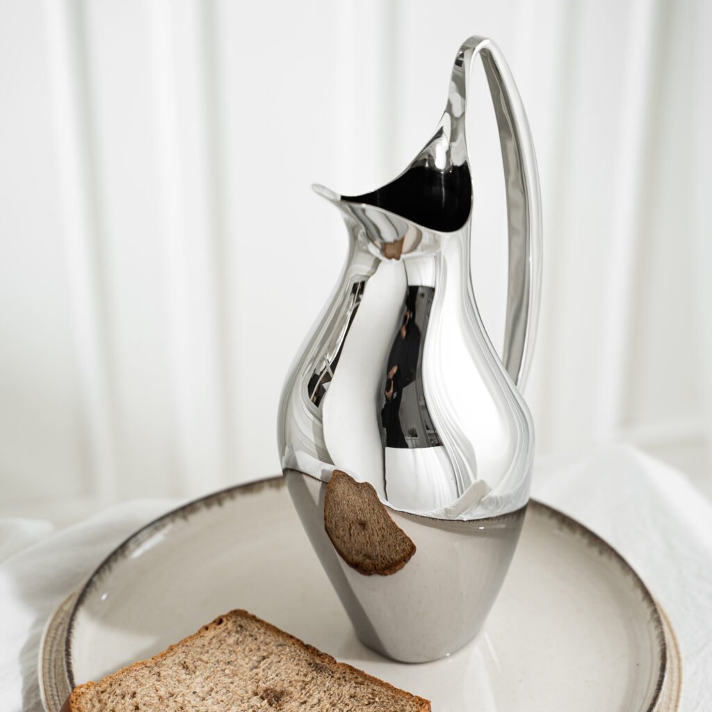 Steel Swan Pitcher placed on a light oak side table beside a minimalist water glass, representing Reflective High-End Kinetic Art elegance.