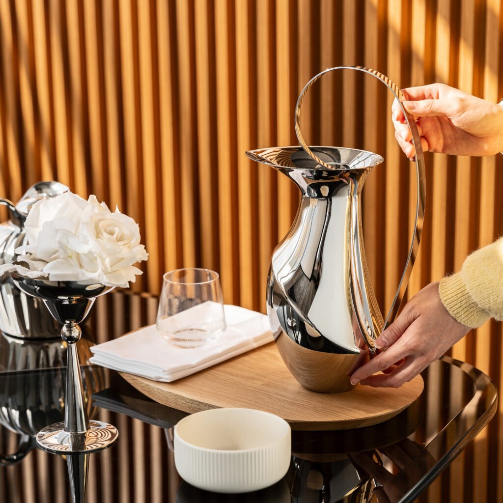 Steel Toucan Pitcher placed on a concrete designer side table beside a glass of sparkling water, representing Reflective High-End Kinetic Art elegance.
