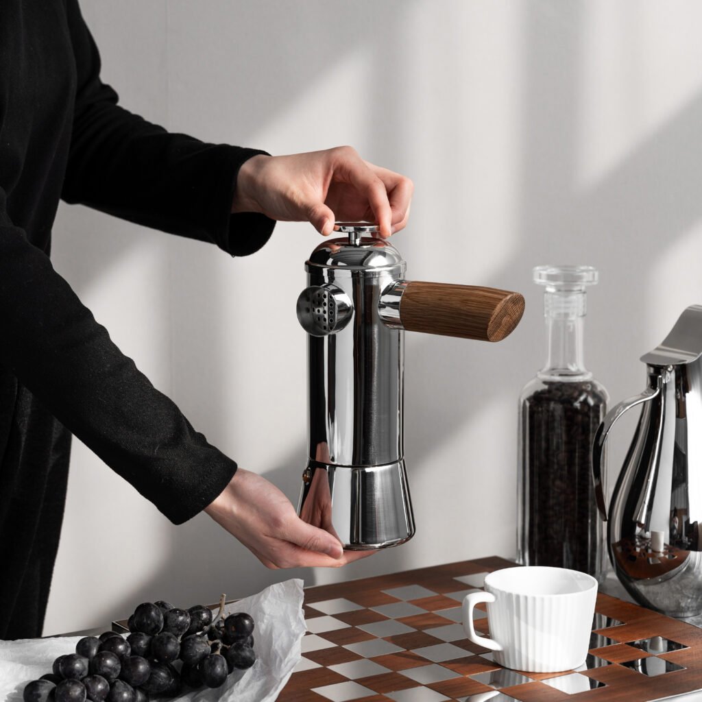 Steel Moka Pitcher placed on a minimalist stone console beside a single tall botanical stem, representing Reflective High-End Vertical Art elegance.
