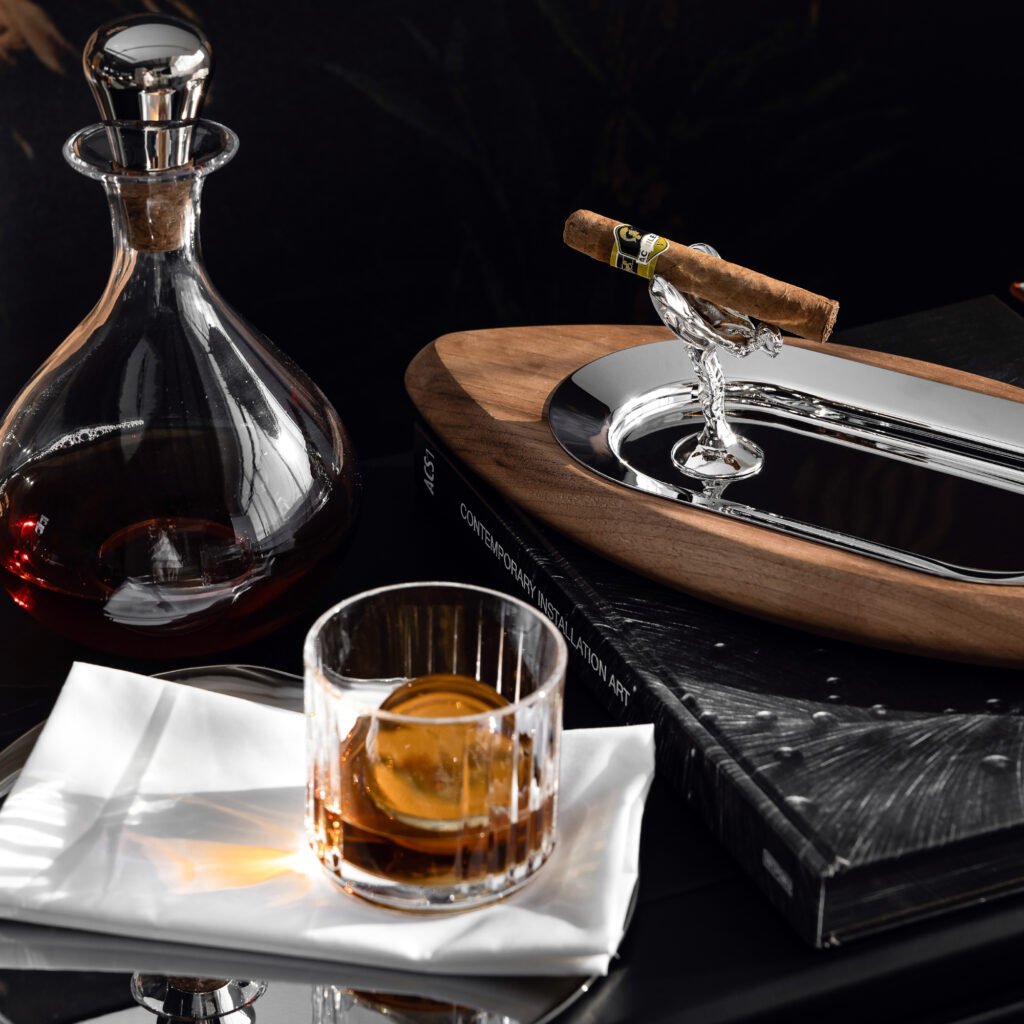 Steel Geometric Ashtray placed on a concrete-textured side table beside a glass of whiskey, representing Reflective High-End Industrial Art elegance.