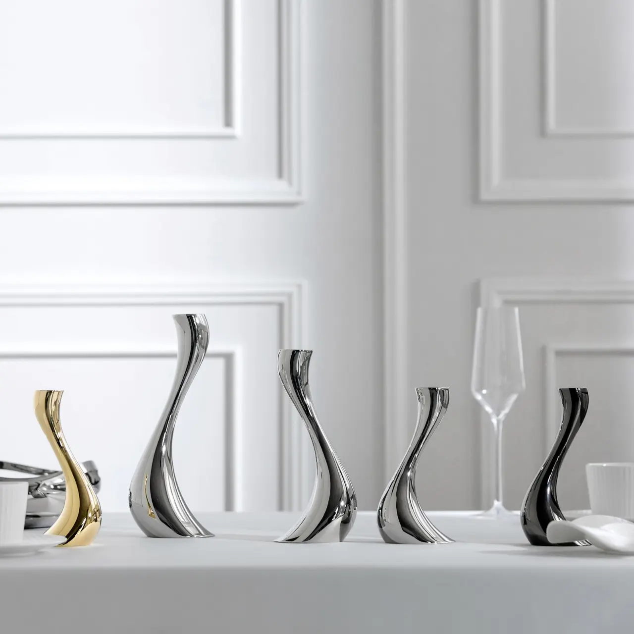 A lineup of stainless steel candle holders in gold and silver mirror finishes displaying various sizes against a clean white wall.