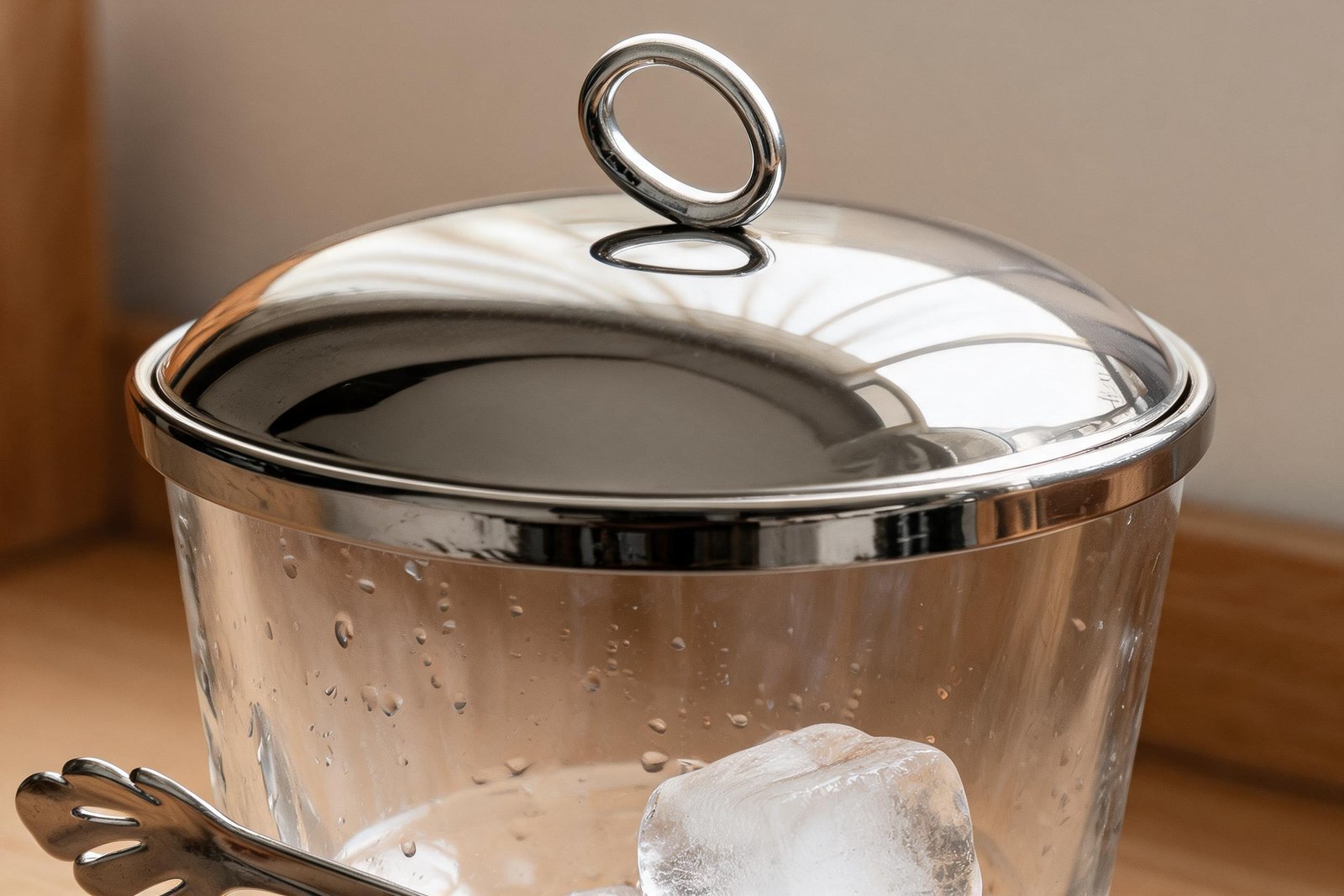 Ice bucket with lid detail shot: the mirror polished stainless steel domed lid and ring handle sitting on the clear glass rim with the steel tongs below