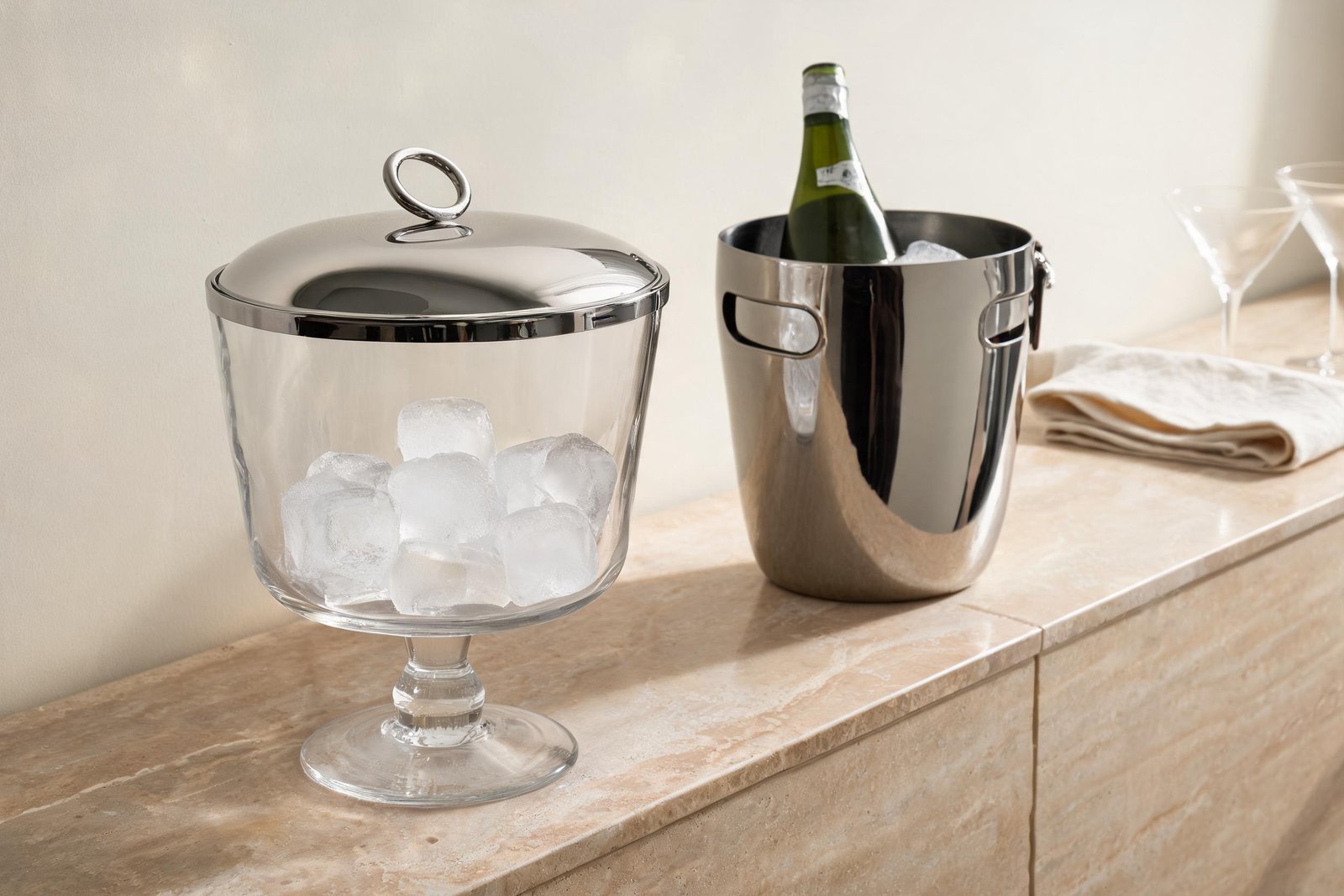 Glass Ice Bucket vs Stainless Steel: What Changes? 4 Glass ice bucket with mirror lid beside an open mirror steel ice bucket on a travertine sideboard, showing the difference in mouth width, lid and stem